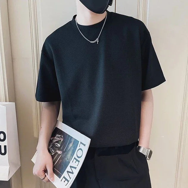 Man wearing a black short-sleeve shirt with a necklace, holding a magazine, in a casual setting.